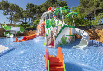 Parco giochi acquatico per bambini con scivoli, palme e spruzzi vicino a una casa mobile sotto il sole.
