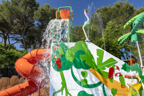 Aire de jeux aquatique avec toboggan orange et décor tropical dans un camping glamping entouré d’arbres.