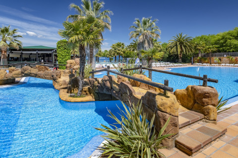Piscine extérieure avec palmiers et pont en pierre au Camping Solmar - XALET, Espagne, sous un ciel bleu clair.