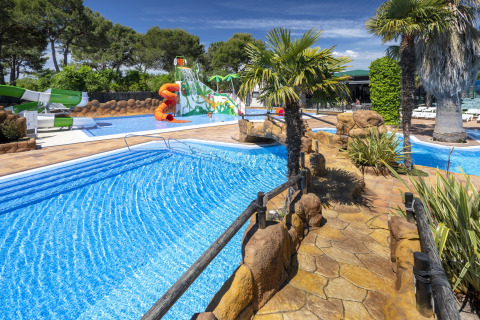 Espace piscine extérieure dans un glamping, avec toboggans, palmiers, sentiers en pierre et aire de jeux.