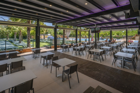 Area pranzo coperta all’aperto in un glamping, con tavoli, sedie e vista su piscina e palme.