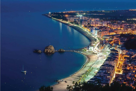 Vue nocturne sur une ville côtière et plage éclairées, depuis un hébergement de glamping en hauteur.