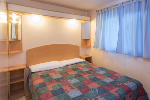 Et soveværelse i mobilhjemmet Camping Solmar - COTTAGE B i Spanien, med farverigt sengetæppe og gardiner.