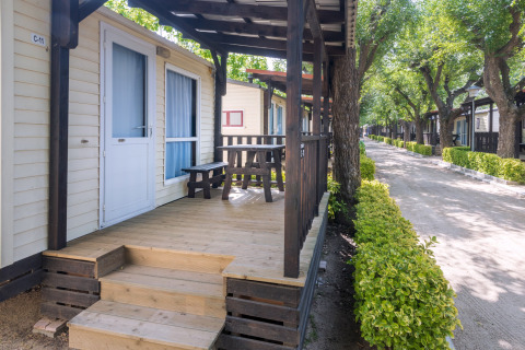 Mobilhome med træveranda og bænk på Camping Solmar i Spanien, omgivet af grønne træer og hække.