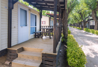 Mobilhome med træveranda og bænk på Camping Solmar i Spanien, omgivet af grønne træer og hække.