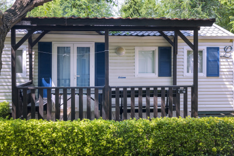 Stacaravan met overdekte veranda en blauwe luiken bij Camping Solmar COTTAGE B, achter groen hegje in Spanje.