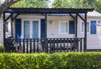 Casa mobile con veranda coperta e persiane blu al Camping Solmar COTTAGE B, vista dietro una siepe verde in Spagna.