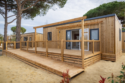 Moderne mobilhome Beach Resort Agde 2 med træterrasse, rampe og omgivet af træer og sand.