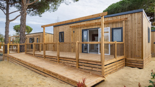 Strandresort Agde 2 Mobilheim mit Holzterrasse und Rampe, umgeben von Bäumen und Sand.
