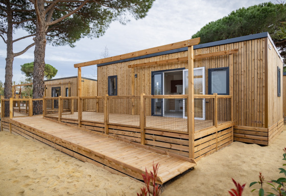 Moderne mobilhome Beach Resort Agde 2 med træterrasse, rampe og omgivet af træer og sand.