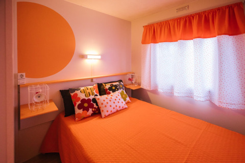 Lichte slaapkamer in een Spaanse stacaravan met oranje beddengoed, kleurrijke kussens en stipgordijnen.