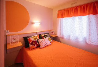Lichte slaapkamer in een Spaanse stacaravan met oranje beddengoed, kleurrijke kussens en stipgordijnen.