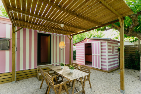 Buiten eethoek met houten tafel en stoelen onder pergola naast roze-wit gestreepte stacaravans in Spanje.