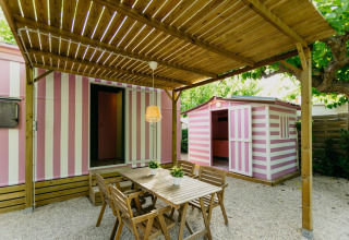 Buiten eethoek met houten tafel en stoelen onder pergola naast roze-wit gestreepte stacaravans in Spanje.