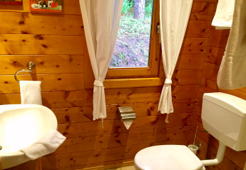 Badeværelse med trævægge, hvidt toilet og håndvask, vindue med gardiner i chalet Remschweiler, Frankrig.