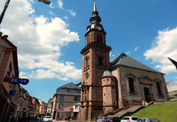 Foto af en historisk kirke i Remschweiler, Frankrig, med blå himmel og skyer i baggrunden.