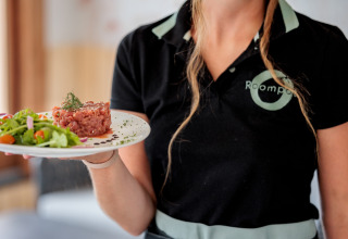 En tjener på glamping-ophold serverer frisk tatar og salat på en restaurant, med Roompot-uniform.