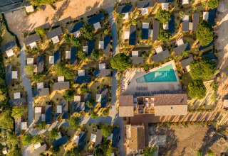 Luftfoto af et glamping-resort med små hytter spredt blandt træer og en stor swimmingpool i midten.