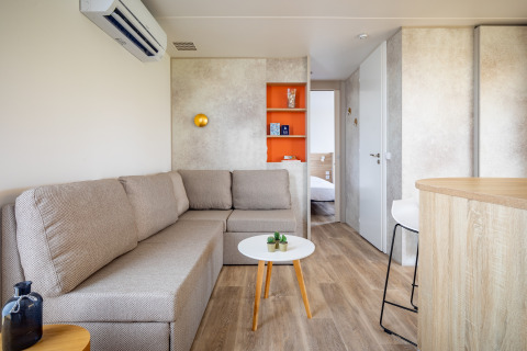 Lyst mobilhjems stue med beige sofa, bord og orange reol på Beach Resort Agde 4 i Frankrig.