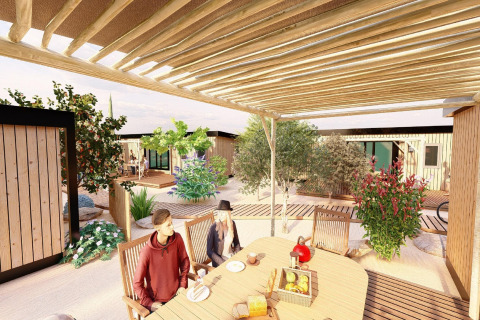 To mennesker sidder ved et bord under en pergola i et mobilhome-resort med træhytter og haveanlæg.