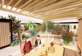 To mennesker sidder ved et bord under en pergola i et mobilhome-resort med træhytter og haveanlæg.