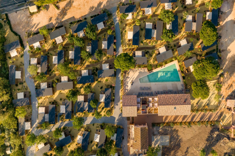 Luftfoto af mobilhome-park med mange små hytter, træer og en stor swimmingpool i centrum.