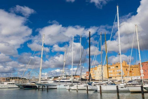 Både fortøjet i en marina med farverige bygninger i baggrunden og en blå himmel med skyer.
