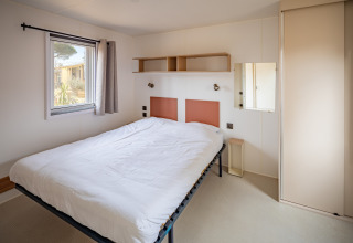 Soveværelse i mobilhome Beach Resort Agde 2 i Frankrig med dobbeltseng, vindue, hylder og skab.
