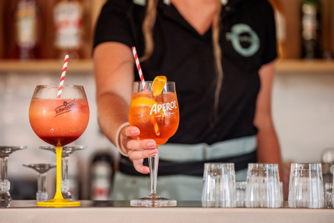 En bartender serverer farverige cocktails med sugerør i store glas foran et barområde i en mobilhome.