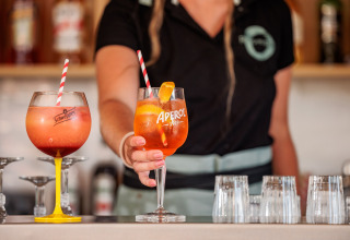 En bartender serverer farverige cocktails med sugerør i store glas foran et barområde i en mobilhome.
