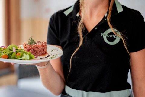 En servitrice i sort Roompot-uniform serverer en tallerken med tartar og salat på Beach Resort Agde 1 i Frankrig.