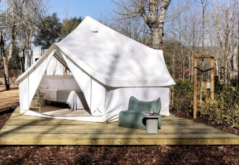 Tenda safari Bell Tent Plus con tela bianca, veranda in legno e arredo esterno tra alberi.