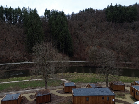 Blick auf das Ferienpark Hameau de la Semois mit Holzhütten, Fluss und bewaldeten Hügeln in Belgien.