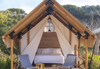 Tenda safari con letto matrimoniale e due sedie su veranda immersa nella natura a Wecamp Santa Cristina, Spagna.