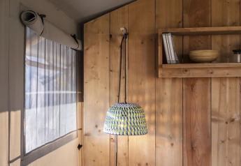 Angolo di una tenda safari con parete in legno, lampada sospesa, finestra a rete e mensola.