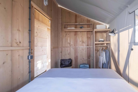 Binnenkant van een safaritent met houten wanden, een bed, een koffer, planken en hangende kleding.
