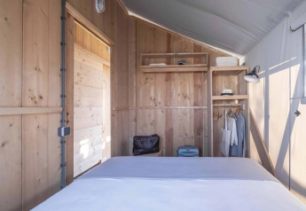 Interno di una tenda safari con pareti in legno, letto, scaffali, valigia e vestiti appesi.