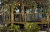 Photo of a wooden lodge cabin nestled among trees, with outdoor shower in the foreground and leafy surroundings.