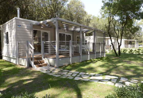 Modern mobiel huis met ruime, overdekte veranda, omgeven door bomen en zonovergoten grasveld.