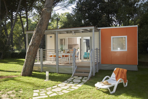 Moderne glampingaccommodatie met veranda, ligstoel en oranje wandpanelen in een groene bosrijke omgeving.