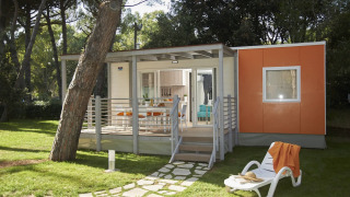 Modernes Glamping-Unterkunft mit Veranda, Sonnenliege und oranger Wand in grüner Waldkulisse.
