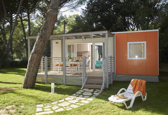 Moderne glampingaccommodatie met veranda, ligstoel en oranje wandpanelen in een groene bosrijke omgeving.
