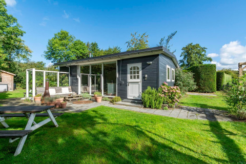 Udendørs foto af lodge Eureka 109 med sort træfacade, stor græsplæne, have og blå himmel.