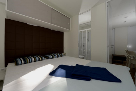 Slaapkamer van een mobilhome met tweepersoonsbed, gestreepte kussens en badkamer zichtbaar door deur.
