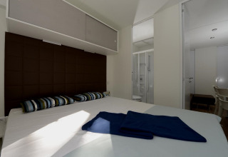 Slaapkamer van een mobilhome met tweepersoonsbed, gestreepte kussens en badkamer zichtbaar door deur.