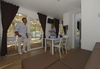 Modern interieur van een mobilhome met twee volwassenen, eettafel, bank en zicht op terras door glazen deuren.