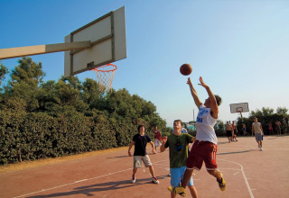 Tieners spelen basketbal op een buitenveld, omgeven door groen en een heldere lucht op een zonnige dag.