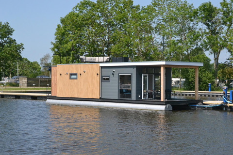 Moderne husbåd ved vandkanten, omgivet af træer, RiggelBrug Sneekermeer, under en blå himmel.