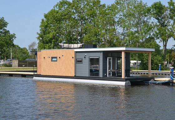 Moderne husbåd ved vandkanten, omgivet af træer, RiggelBrug Sneekermeer, under en blå himmel.
