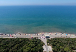 Luftaufnahme eines Strandes mit bunten Sonnenschirmen, Schwimmern und Glamping-Unterkünften am Meer.